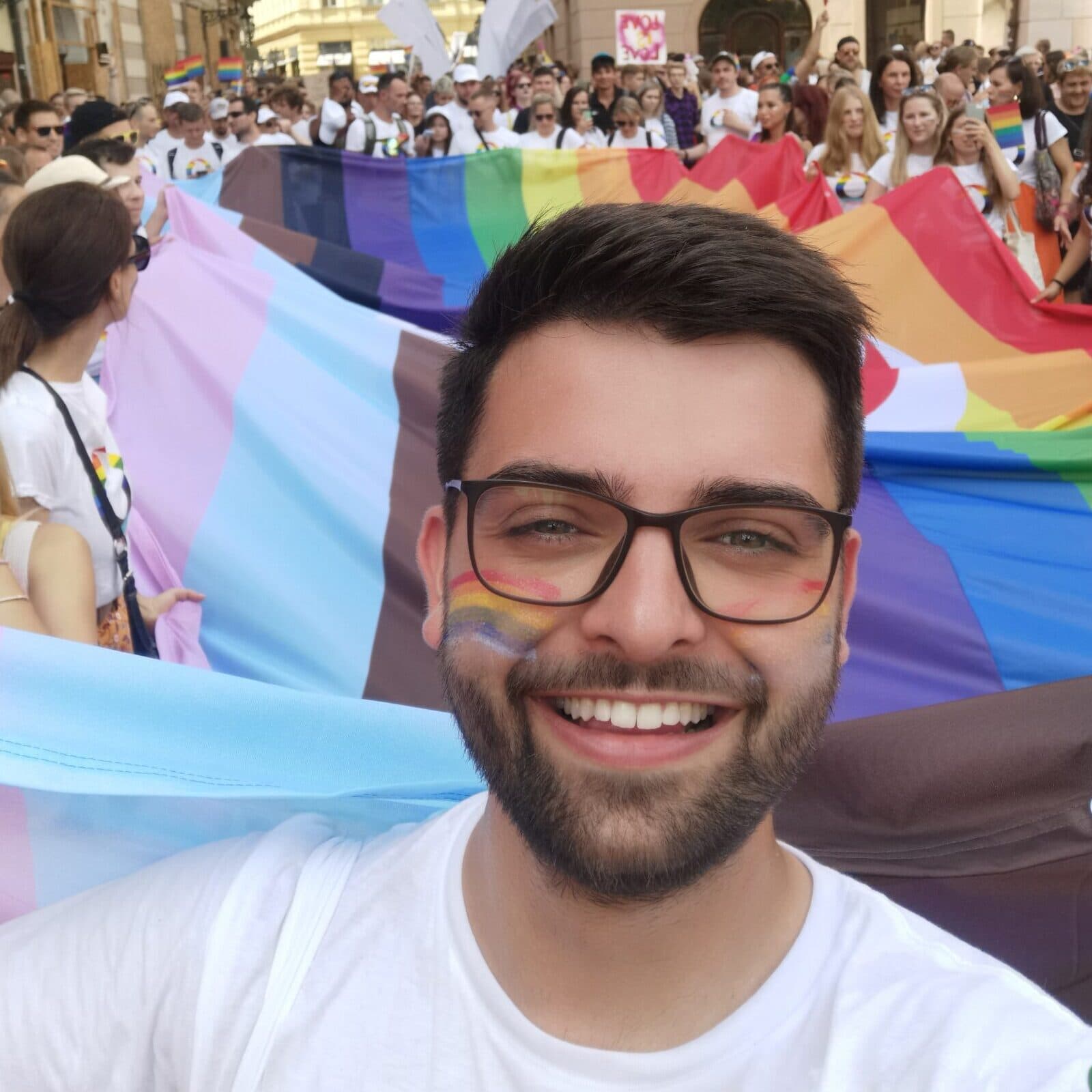 Ondřej smiling in front of a large rainbow flag during a pride parade in Prague.