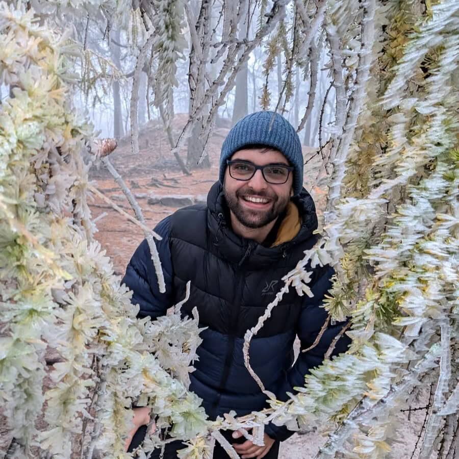 Ondřej smiling in a beanie and glasses, framed by frosty white branches during a winter hike