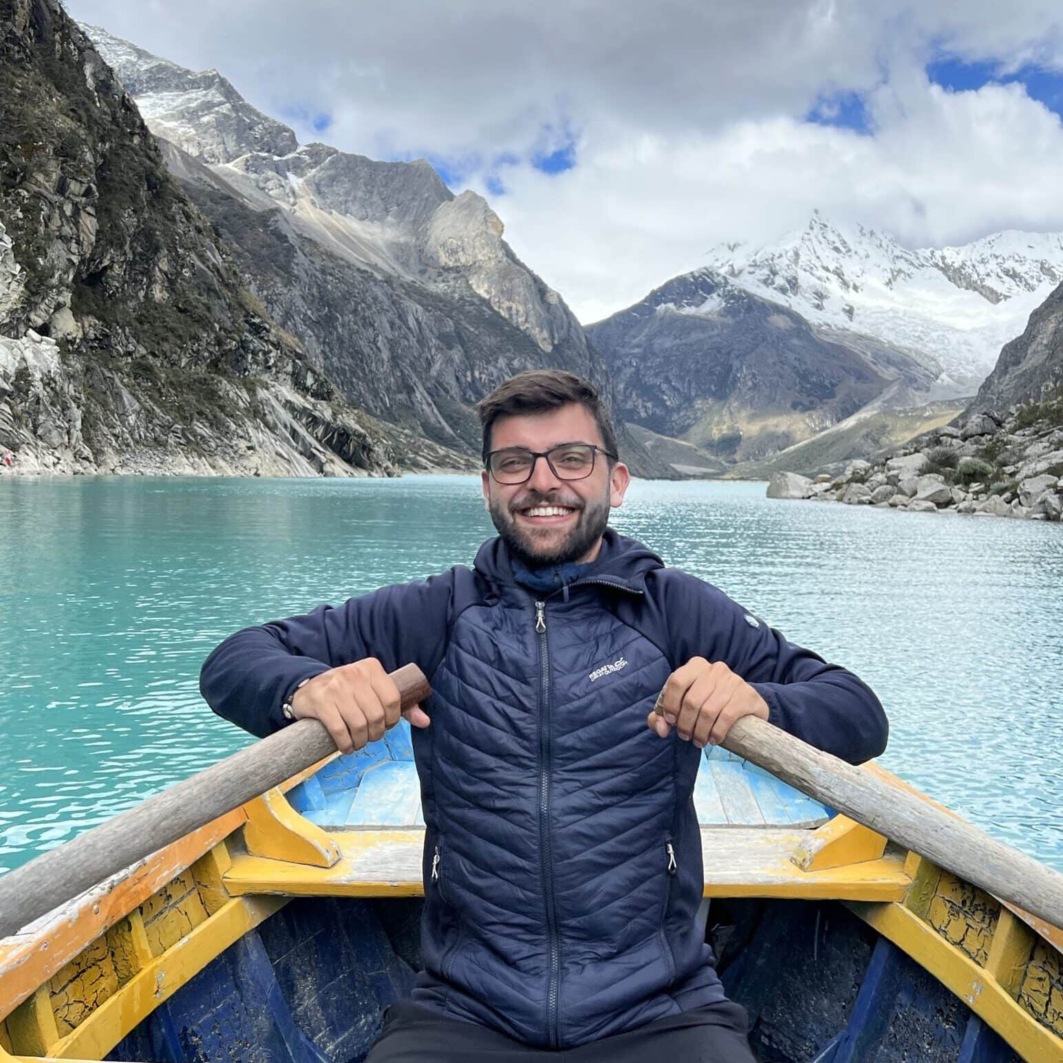 Ondřej, sitting in a yellow wooden boat on a turquoise mountain lake, smiling with snow-covered peaks behind